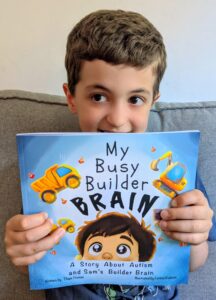 Child holding a copy of My Busy Builder Brain, a children's picture book about autism inspired by his love of construction vehicles.
